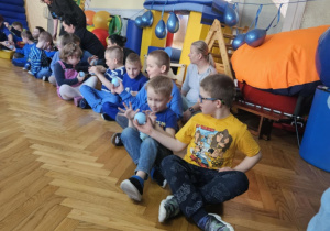 Dzieci siedzą w rzędzie i przekazują sobie niebieską piłkę