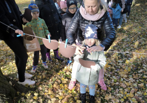 Dziewczynka w niebieskiej czapce zawiesza do sznurka za pomocą klamerki kształt jabłka