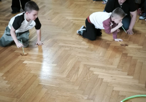 Dwóch chłopców przesuwa po podłodze liść dmuchając poprzez rurkę i wprawiając liść w ruch