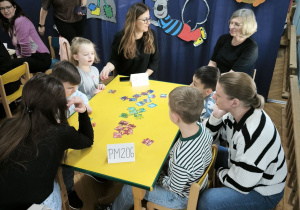 Dzieci z Przedszkola Miejskiego 206 oraz Przedszkola Miejskiego 185 rywalizują w grach planszowych siedząc przy stoliku wraz z opiekunkami