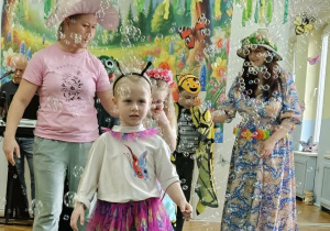Dziewczynka przebrana za motyla spaceruje po zielonym płótnie. Wokół dziecka latają mydlane bańki