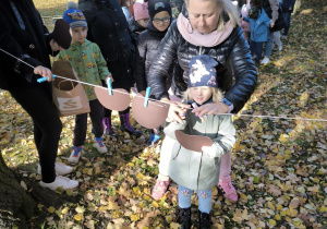 Dziewczynka w niebieskiej czapce zawiesza do sznurka za pomocą klamerki kształt jabłka