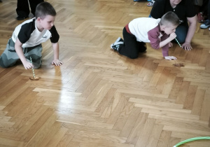 Dwóch chłopców przesuwa po podłodze liść dmuchając poprzez rurkę i wprawiając liść w ruch