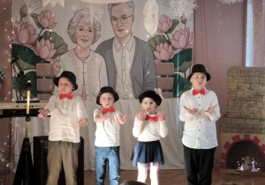 Dzieci z grupy Zajączków występują mając podniesione ręce oraz założona czarne kapelusze