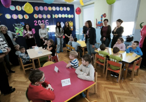 Dzieci z przedszkoli siedzą przy stolikach, czekając na rozpoczęcie olimpiady