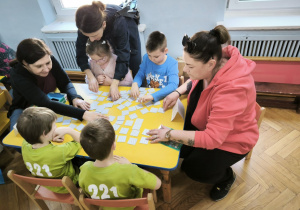 Dzieci z Przedszkola Miejskiego 109 oraz Przedszkola Miejskiego 221 rywalizują w grach planszowych siedząc przy stoliku wraz z opiekunkami
