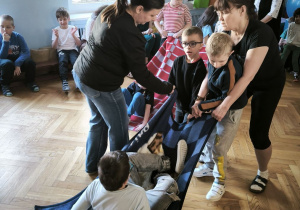 Dwóch chłopców z pomocą nauczycielek przeciąga kolejnego chłopca leżącego na kocu