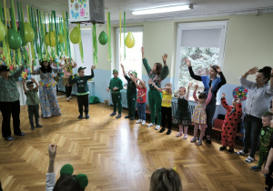 Dzieci i nauczycielki stoją w kręgu i trzymają w górze ręce machając nimi