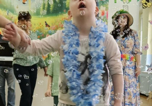 Dziewczynka mająca na szyi biało niebieski wieniec spaceruje po zielonym płótnie. Wokół dziecka latają mydlane bańki