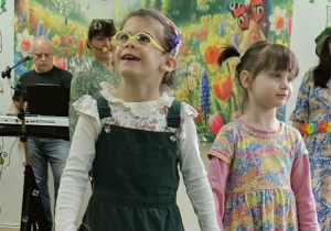 Dziewczynka ubrana w zieloną sukienkę i dziewczynka ubrana w sukienkę w kwiaty spacerują po zielonym płótnie. Wokół dzieci latają mydlane bańki