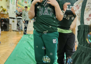 Chłopcy przebrani za żabki spacerują po zielonym płótnie. Wokół dzieci latają mydlane bańki