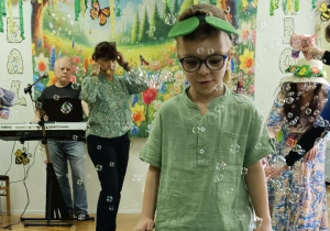 Chłopiec przebrany za żabkę spaceruje po zielonym płótnie. Wokół dziecka latają mydlane bańki