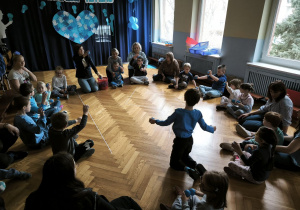 Dzieci siedzą w kręgu i podnoszą do góry dwie, niebieskie piłki