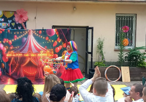 Pani przebrana za clowna trzyma w dłoniach obręcz z siatką i pomarańczowymi piłkami
